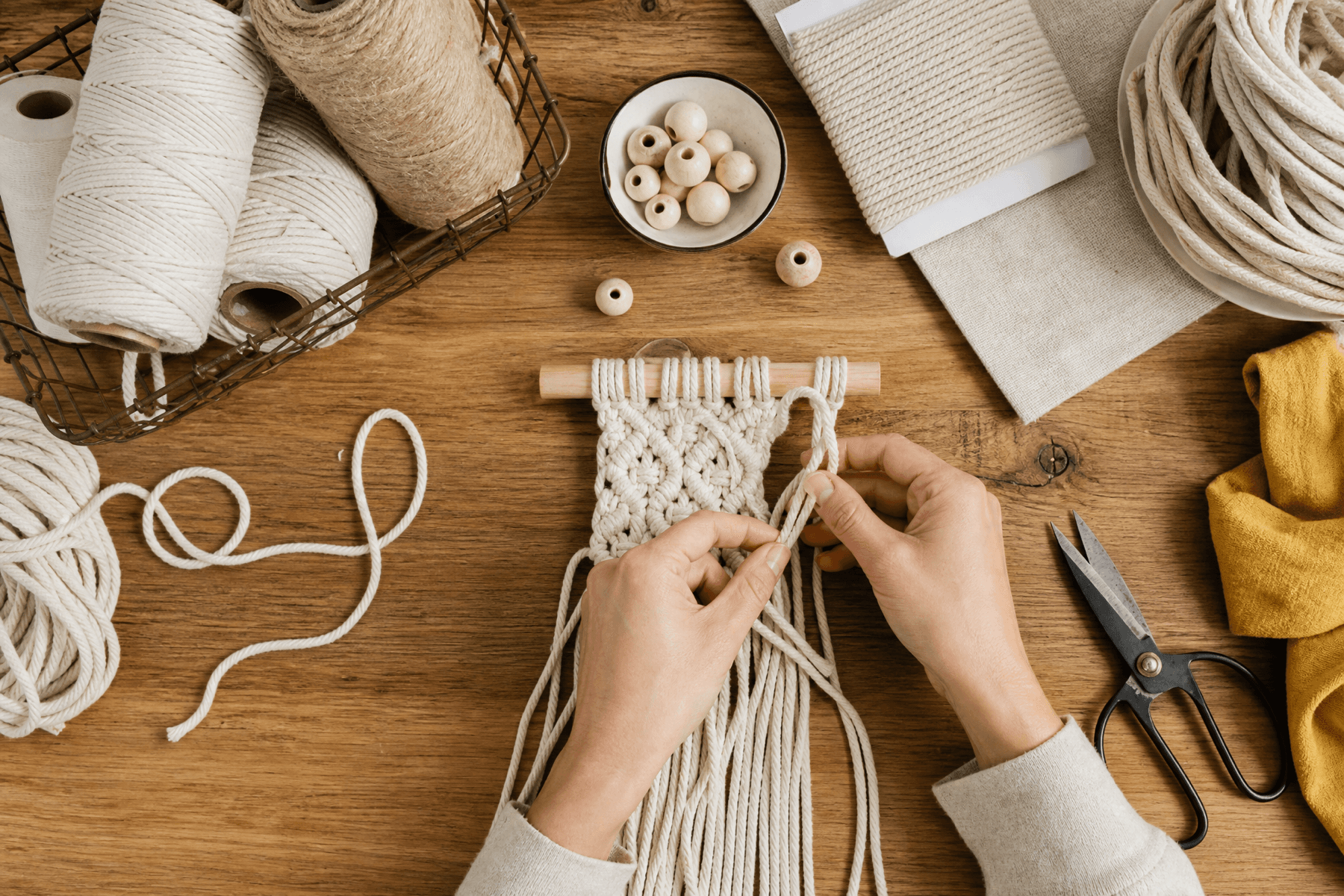 Hands weaving cotton cord on a wooden workbench surrounded by craft materials